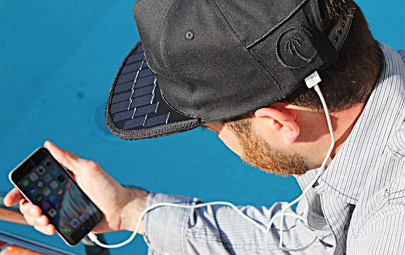 Man wearing a solar-powered cap charging his smartphone via USB while sitting outdoors.